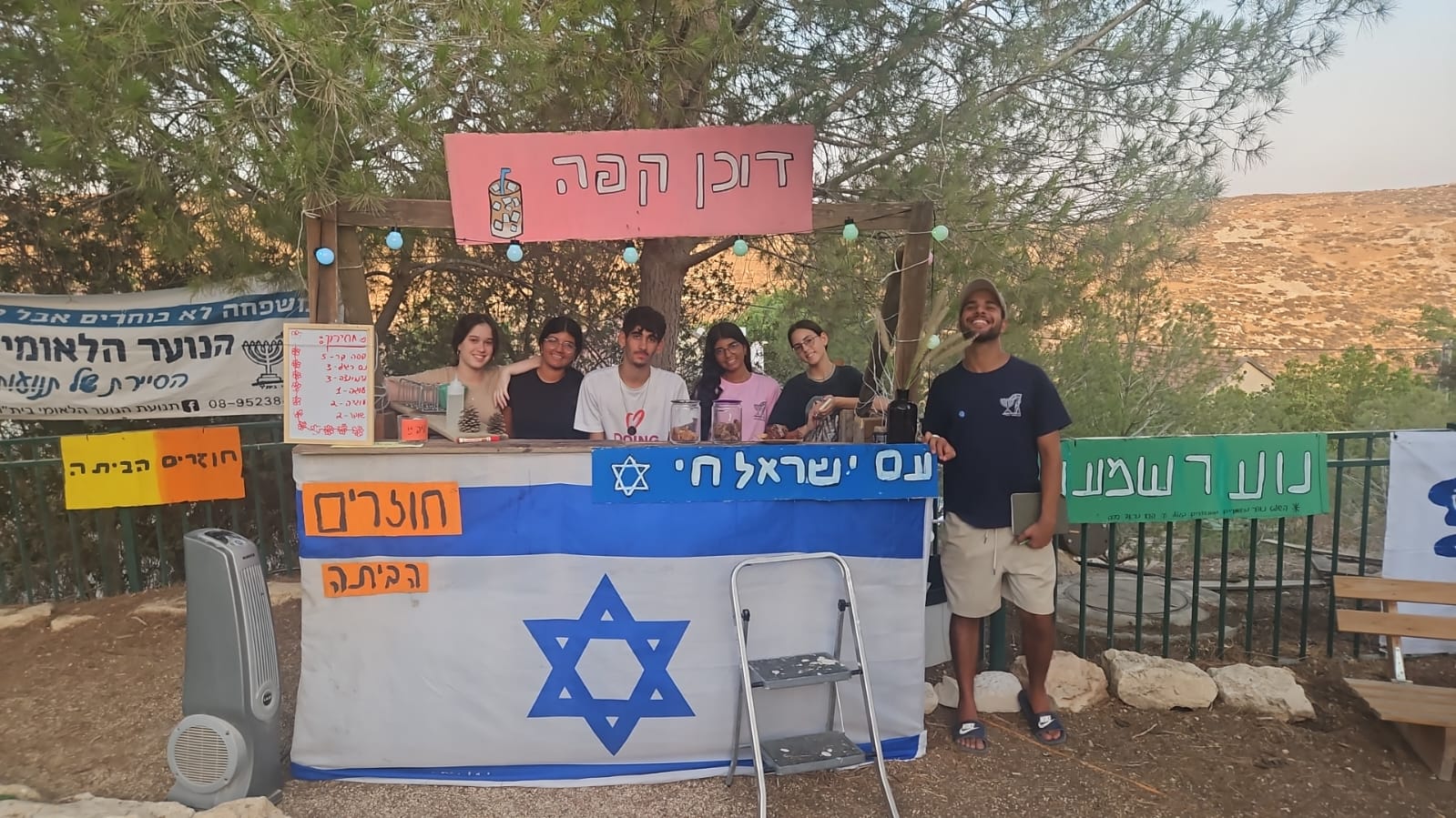 נערים ונערות בשנת שירות של הנוער הלאומי בית"ר בתוך חממה מאחורי דוכן גדול עם דגל ישראל