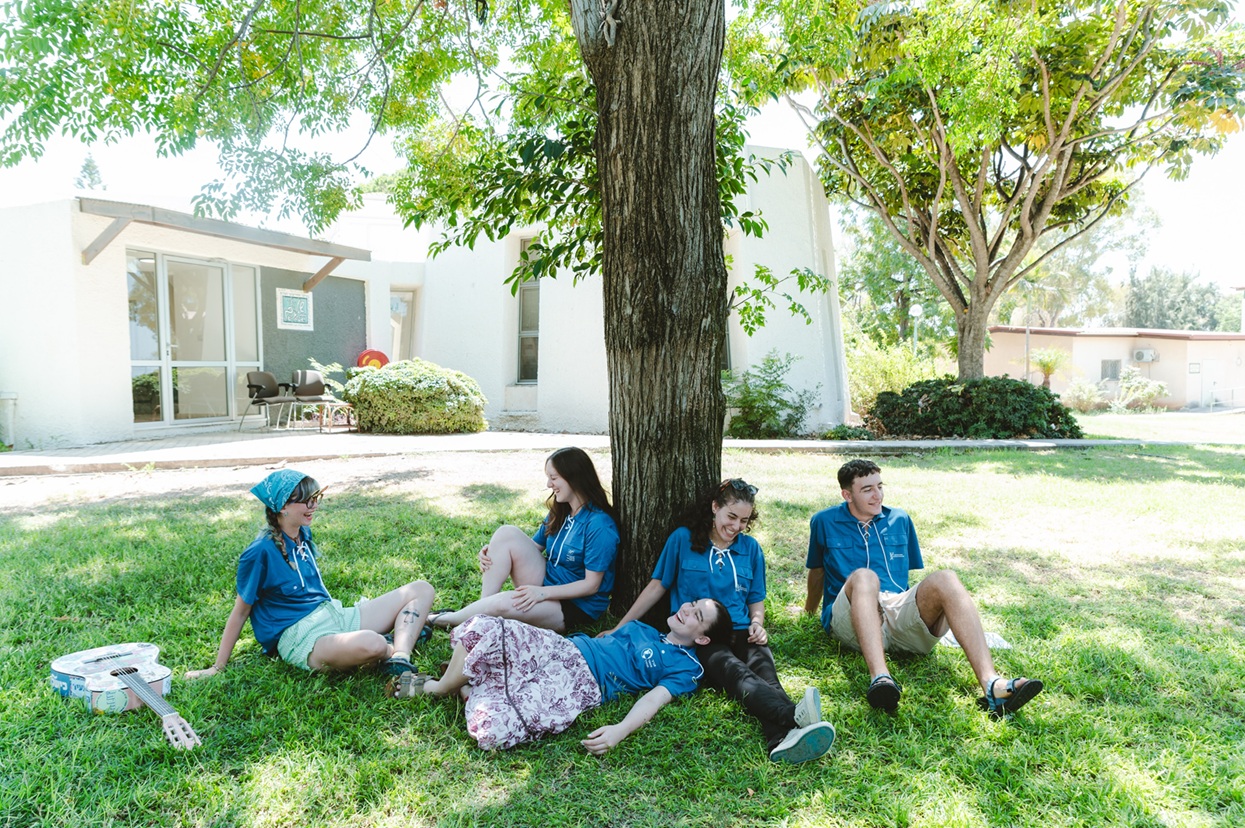 נערים ונערות בחולצות כחולות יושבים על דשא תחת עץ