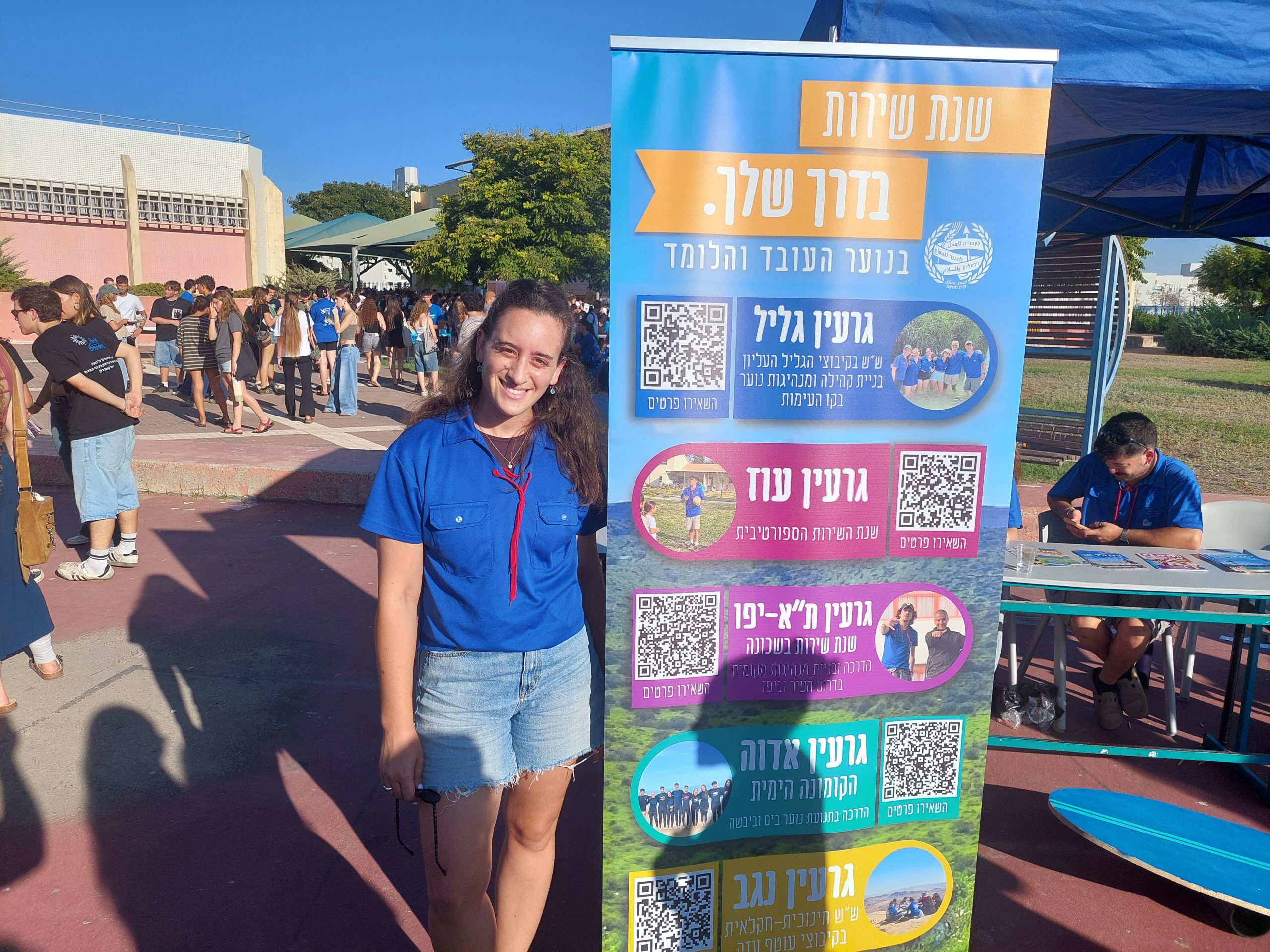 אשה צעירה עם חולצה כחולה עומדת בכנס ליד שלט של הנוער העובד והלומד