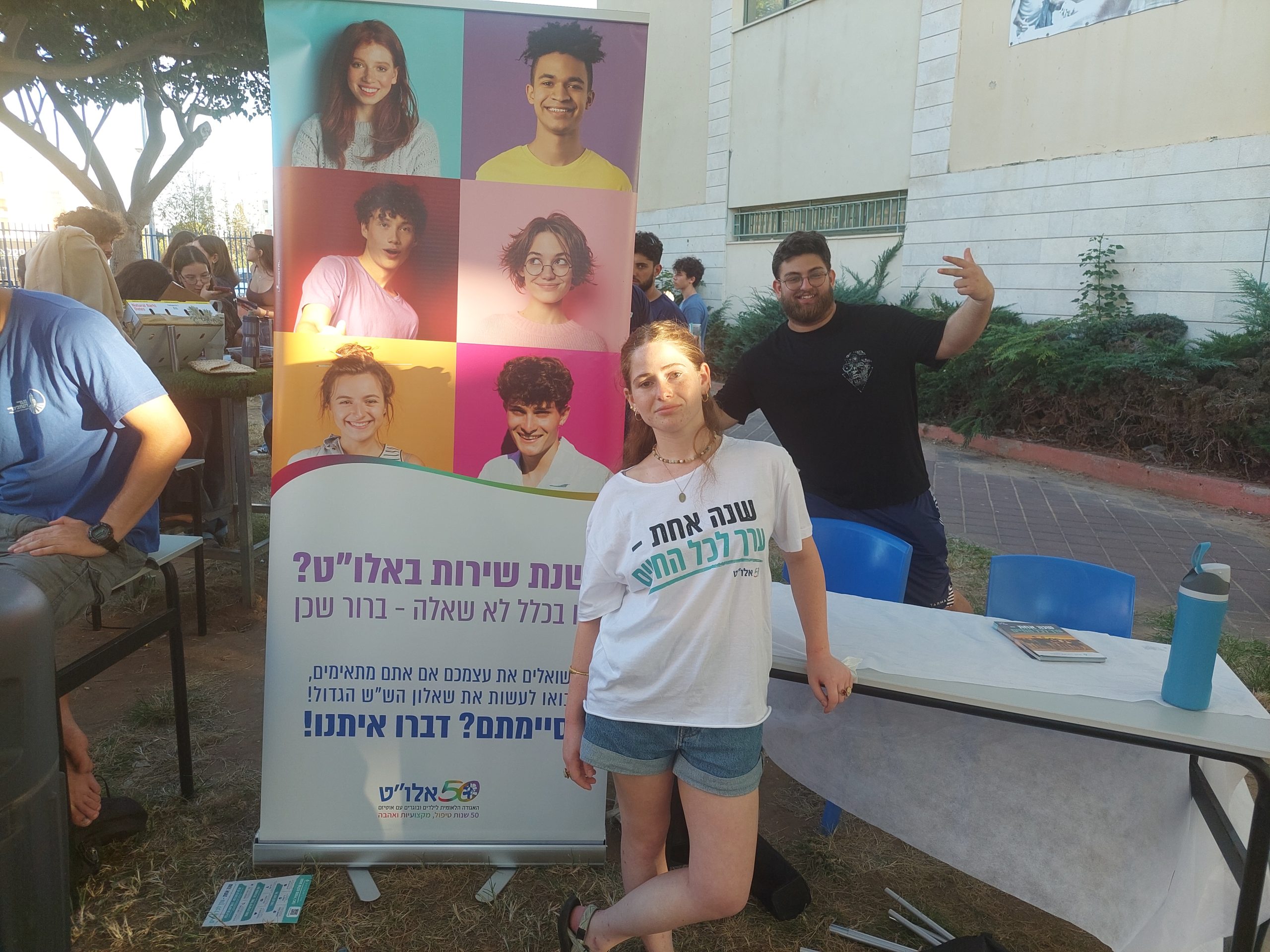 נערה בחולצה לבנה עומדת ליד שלט של עמותת אלו"ט. מאחוריה נער בחולצה שחורה